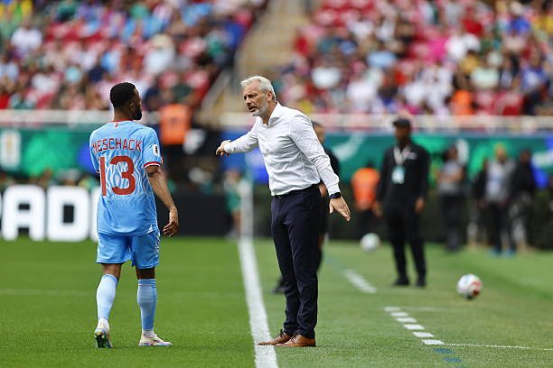 Soccer coach in a white shirt arguing with a player in light blue jersey on the sideline, stadium filled with fans in the background.