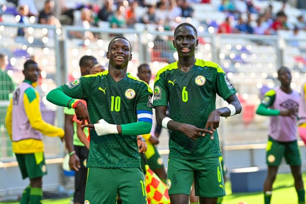 Two Senegalese football players in green kits celebrate on the pitch as fans cheer in the stands behind them.