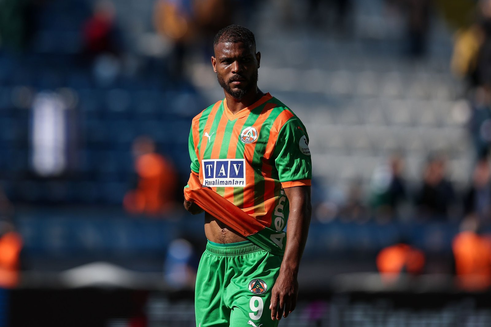 Soccer player in a green and orange striped jersey standing on a stadium field, looking to the side; visible team crest and sponsor logo on chest.