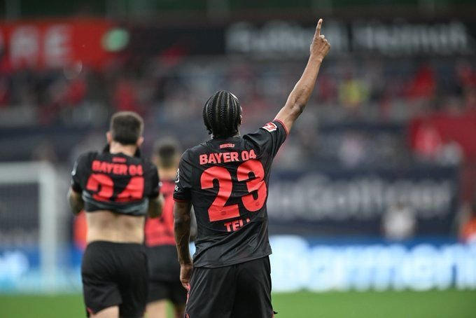 Soccer player in Bayer 04 jersey number 23 raises his right arm during a match, back view.