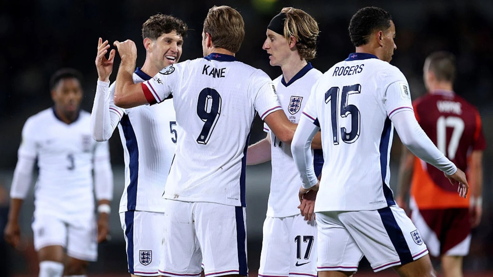 England soccer players celebrate a goal on the field in white kits, smiling and high-fiving each other in a group hug.