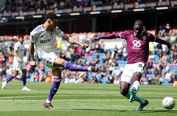 Two soccer players contest the ball on a sunny stadium pitch during a match, one in white kit and purple socks, the other in maroon jersey and white shorts, with spectators in the background.
