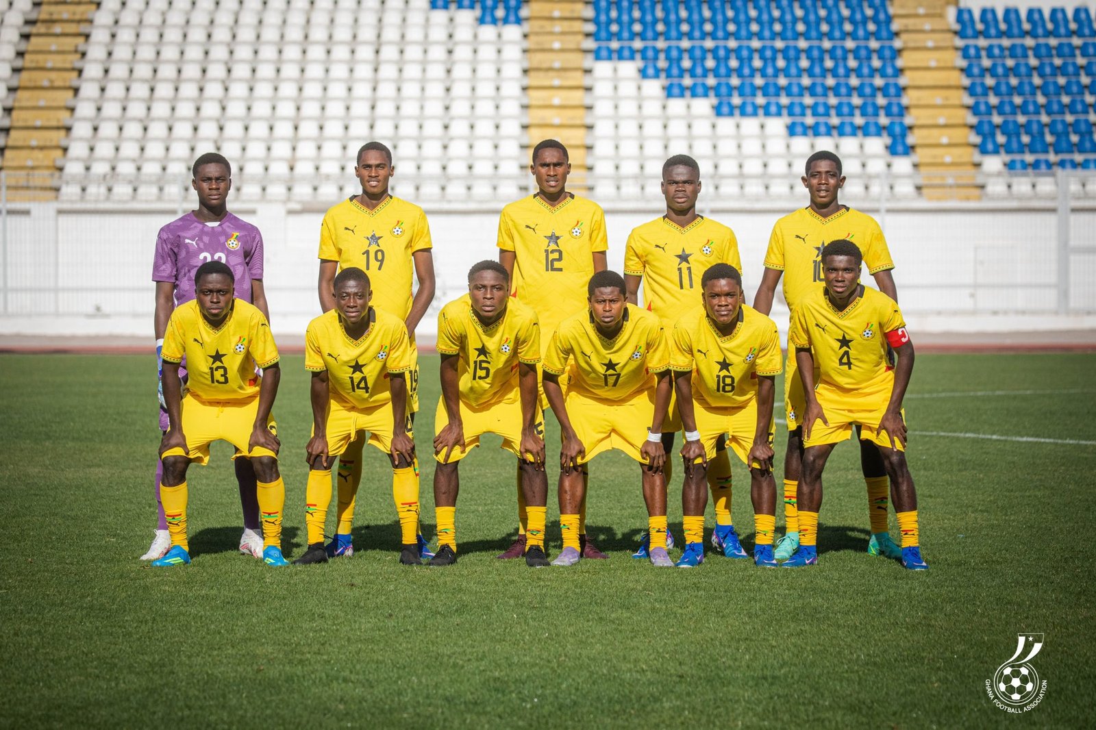 Soccer team in yellow uniforms posing for a group photo on a green field with an empty stadium in the background, goalkeeper in purple on the left. Soccer team in yellow uniforms posing for a group photo on a green field with an empty stadium in the background, goalkeeper in purple on the left.