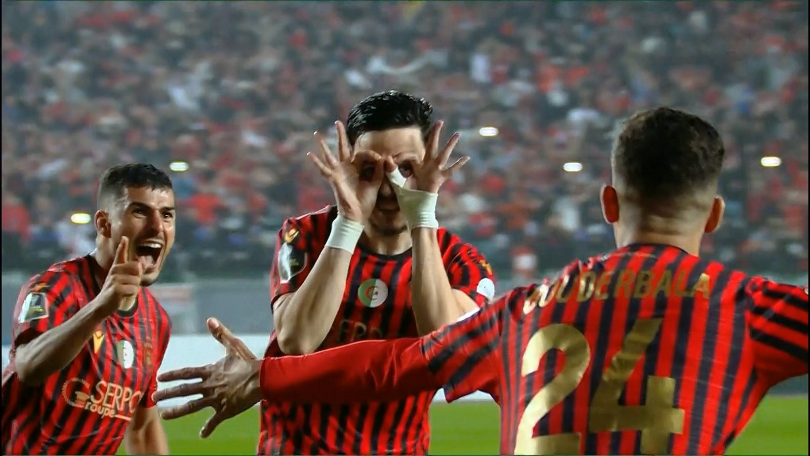 Soccer players in red-and-black striped uniforms celebrate on the field with a cheering crowd in the background, central player making an eye gesture with hands. Soccer players in red-and-black striped uniforms celebrate on the field with a cheering crowd in the background, central player making an eye gesture with hands.