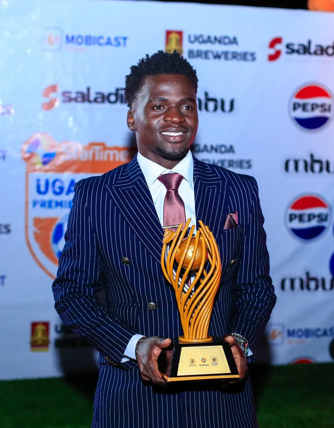 Man in a navy pinstripe suit holding a golden trophy at an awards event, smiling for the camera with sponsor banners in the background.