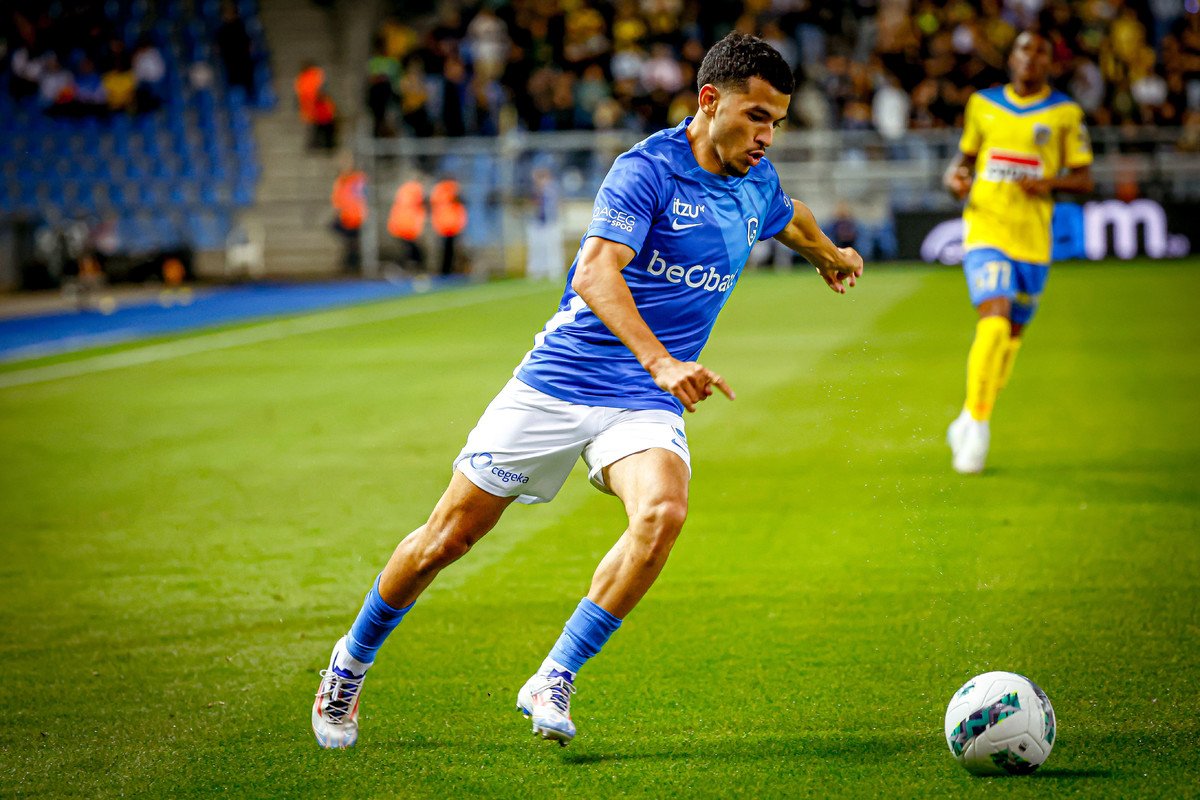 Soccer player in blue jersey dribbling a ball on green field with a yellow-clad opponent in the background.