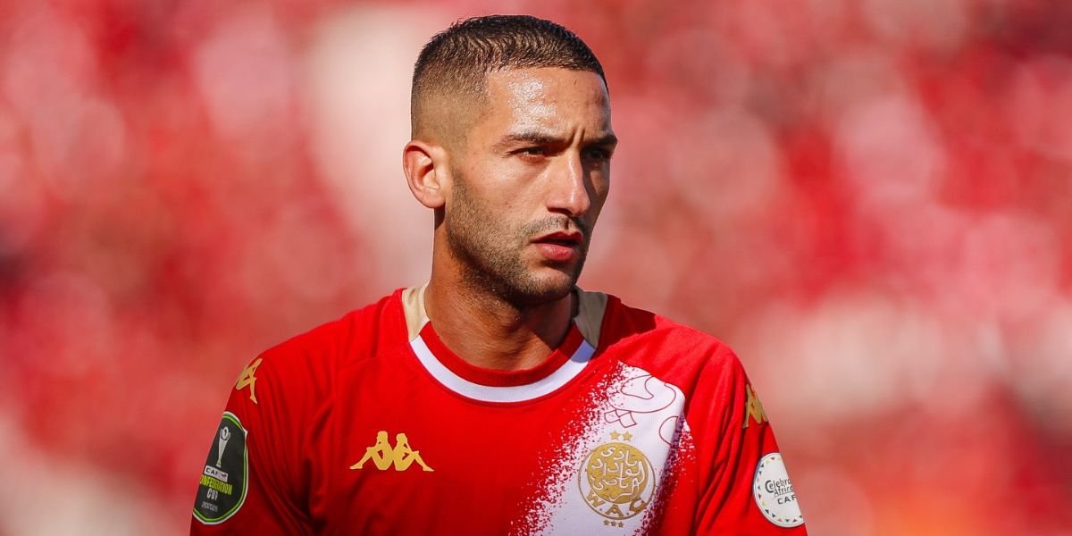Male soccer player in a red jersey with gold logos, looking to the left with a blurred stadium crowd in the background.
