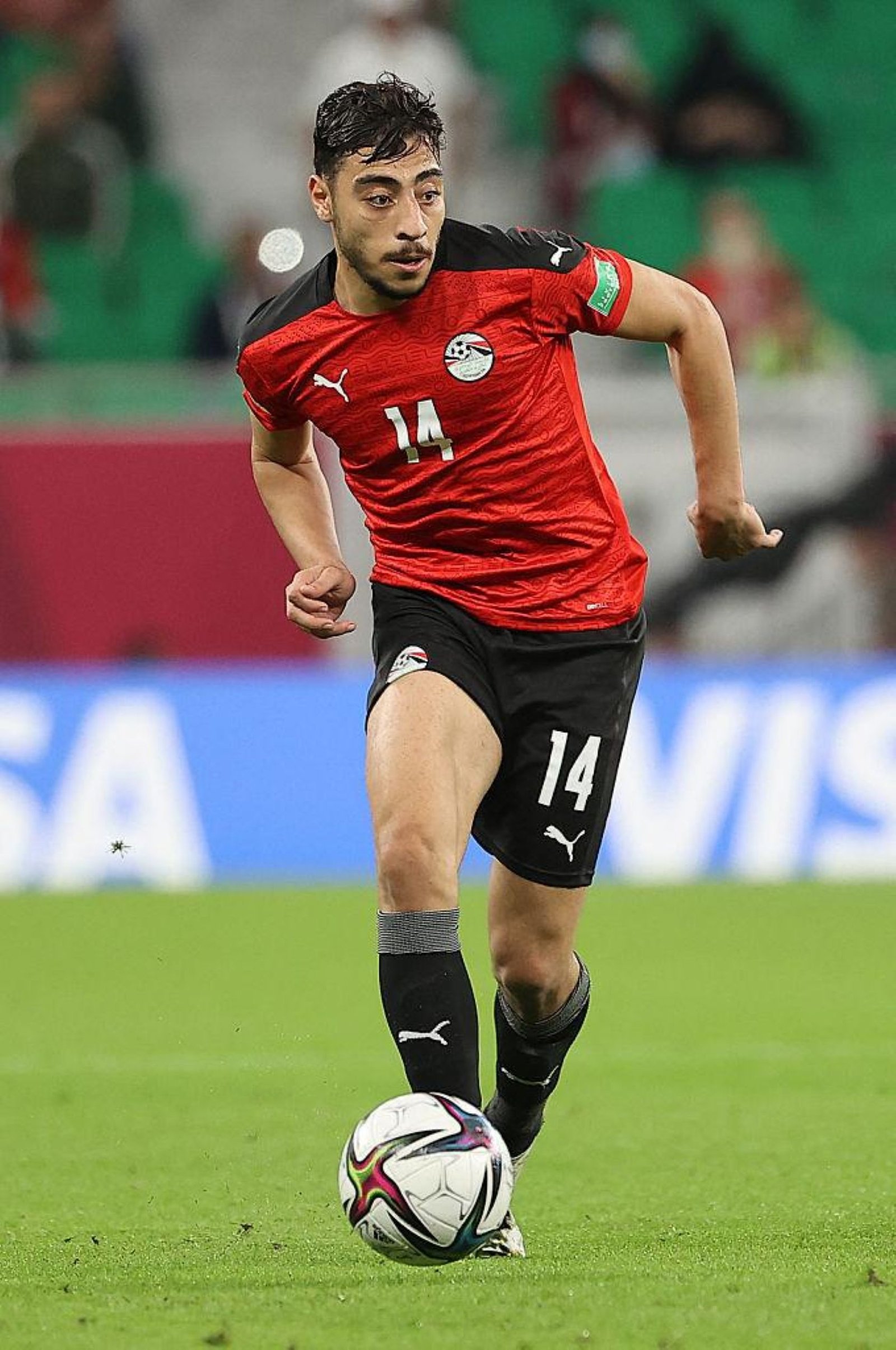Soccer player in a red jersey with number 14 dribbling a ball on a green pitch during a match. Soccer player in a red jersey with number 14 dribbling a ball on a green pitch during a match.