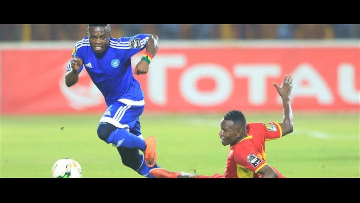 Soccer player in blue uniform dribbles the ball as a red-uniform defender challenges him on a grassy field in motion.
