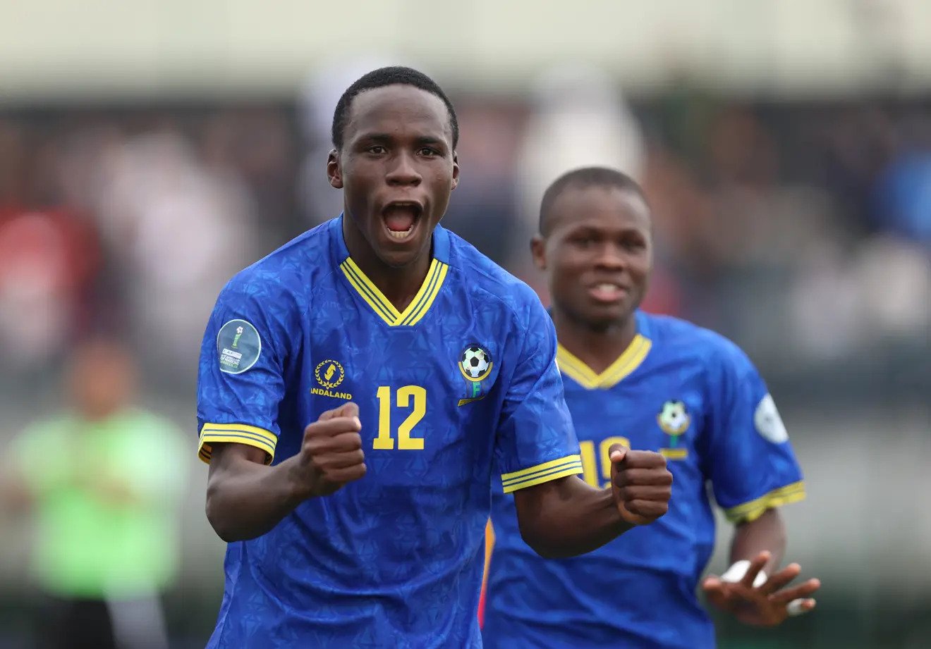 Two soccer players in blue uniforms celebrate with fists raised on the field, one forward shouting in triumph.