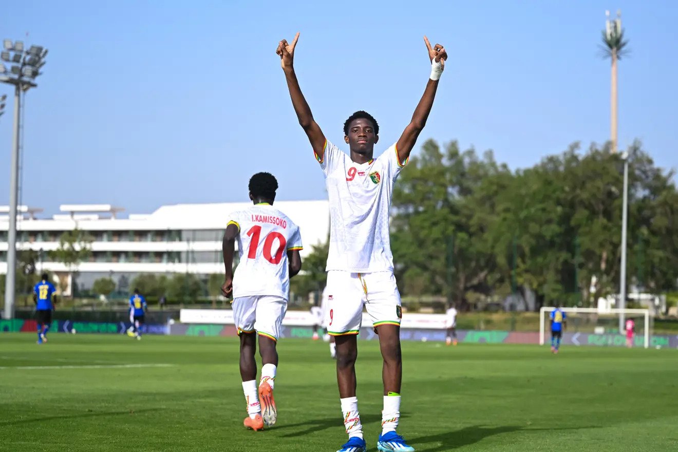 Soccer player in a white jersey with the number 9 raises both arms in celebration on a bright green pitch, with a teammate wearing number 10 in the background.