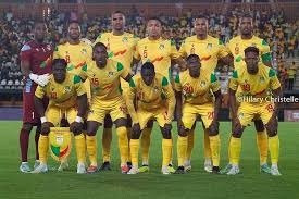 Nine football players in yellow uniforms pose for a team photo on the field before a match, with spectators in the stands behind them.