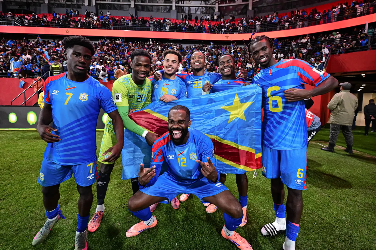 Six soccer players in blue uniforms celebrate on the field, holding a blue flag with a yellow star and diagonal stripe behind them, stadium crowd in the background. Six soccer players in blue uniforms celebrate on the field, holding a blue flag with a yellow star and diagonal stripe behind them, stadium crowd in the background.