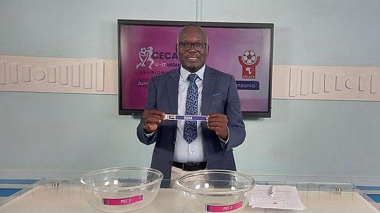 Man in a blue suit holds a lottery ticket while standing behind a table with two clear bowls; a monitor with lottery branding is behind him.