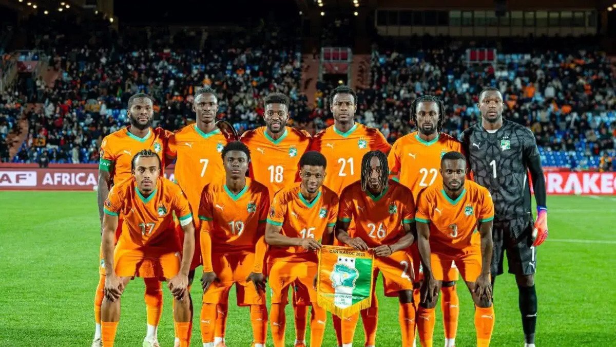 Group of soccer players in orange kits posing for a team photo on a green pitch with a packed stadium in the background, some players kneeling in front and others standing, one player holding a banner. Group of soccer players in orange kits posing for a team photo on a green pitch with a packed stadium in the background, some players kneeling in front and others standing, one player holding a banner.