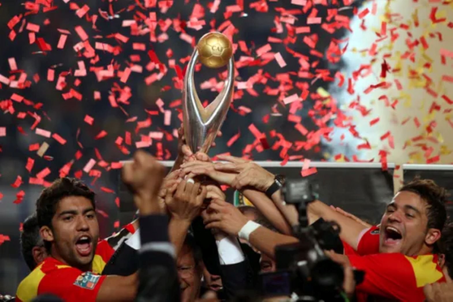 Athletes raise a trophy aloft as red confetti falls during a victory celebration while fans cheer nearby.