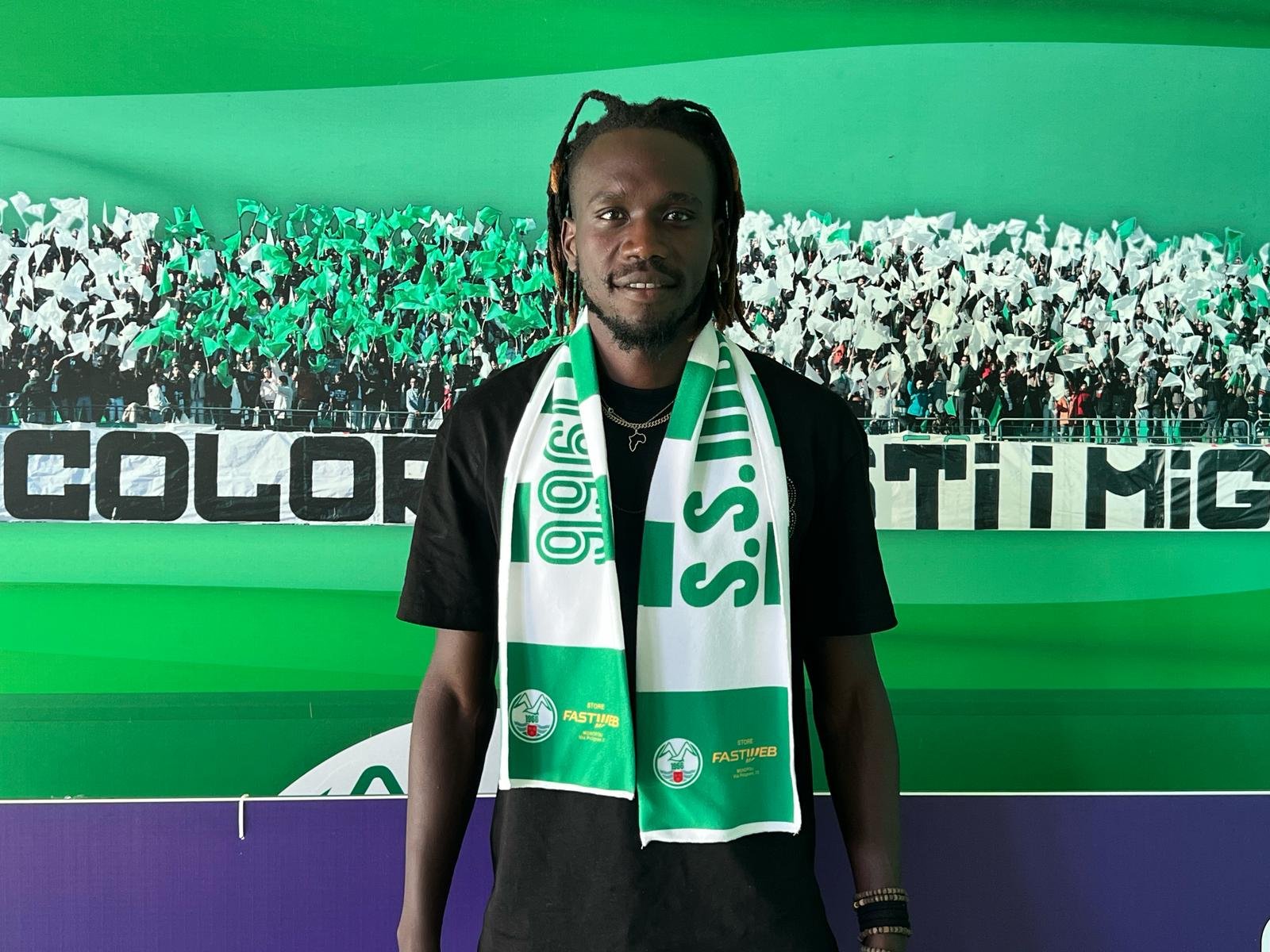 Portrait of a man in a black shirt wearing a green-and-white scarf, standing in front of a packed stadium crowd with green banners behind him.