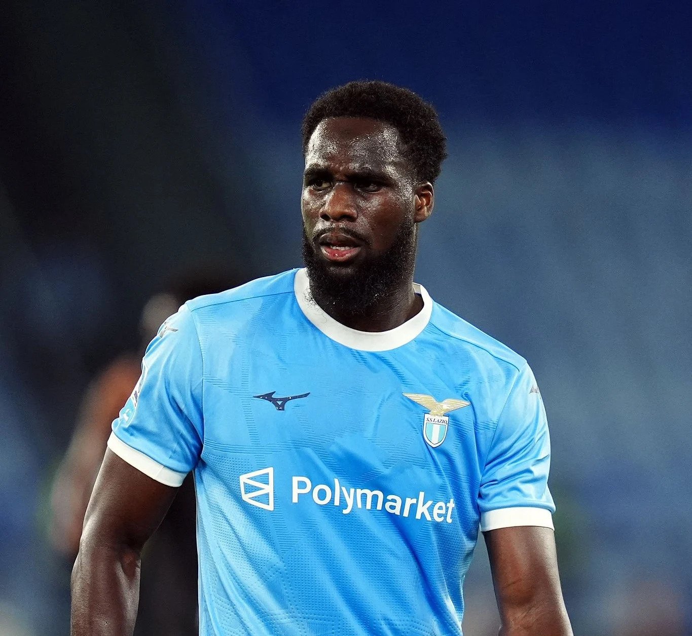 Dark-skinned footballer in a light-blue Lazio jersey on the pitch, looking to the left with a focused expression, sweat visible on his face. Dark-skinned footballer in a light-blue Lazio jersey on the pitch, looking to the left with a focused expression, sweat visible on his face.