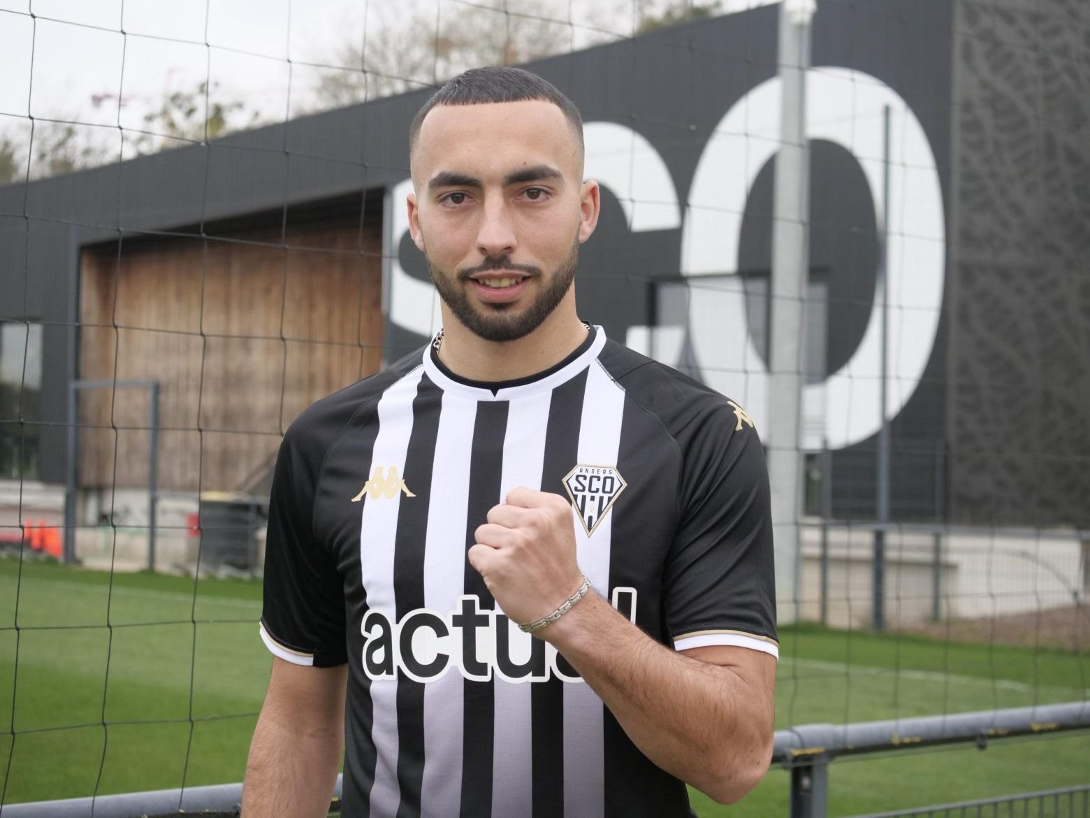 Soccer player in black-and-white striped jersey making a fist toward the camera on a sports field behind a fence recession.