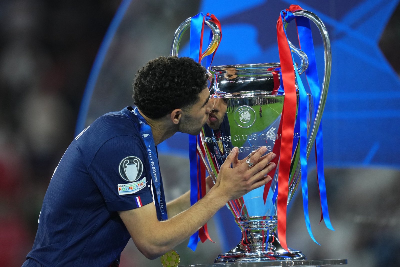 Soccer player in a blue jersey kissing a large silver trophy decorated with red and blue ribbons. Soccer player in a blue jersey kissing a large silver trophy decorated with red and blue ribbons.