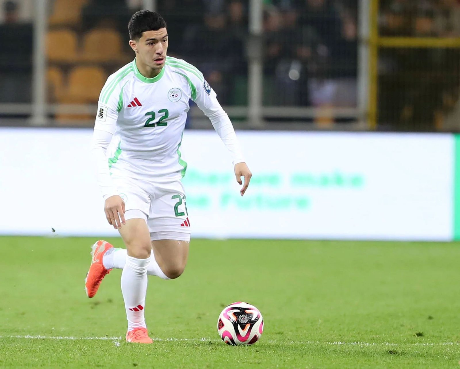 Soccer player in white and green kit (number 22) dribbling a ball on a green field during a match, stadium in background.