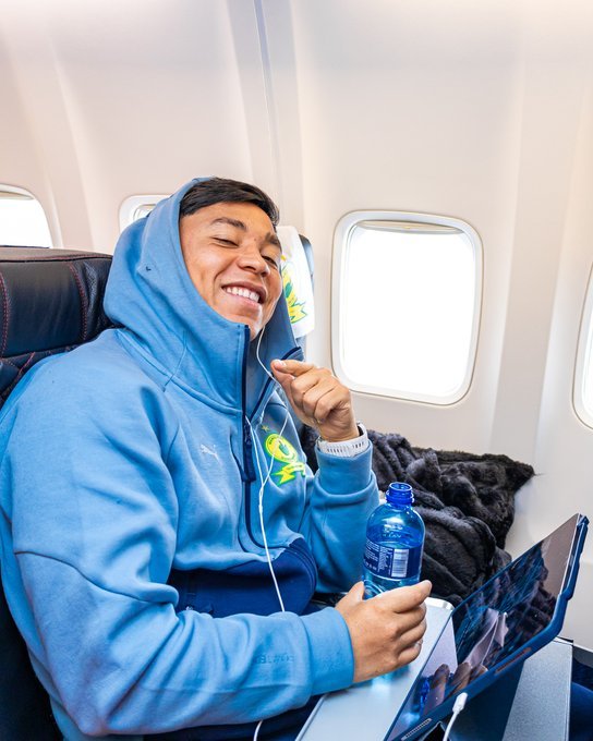 Young man in a blue hoodie on a plane, smiling with earbuds in, holding a water bottle and laptop on the tray table.