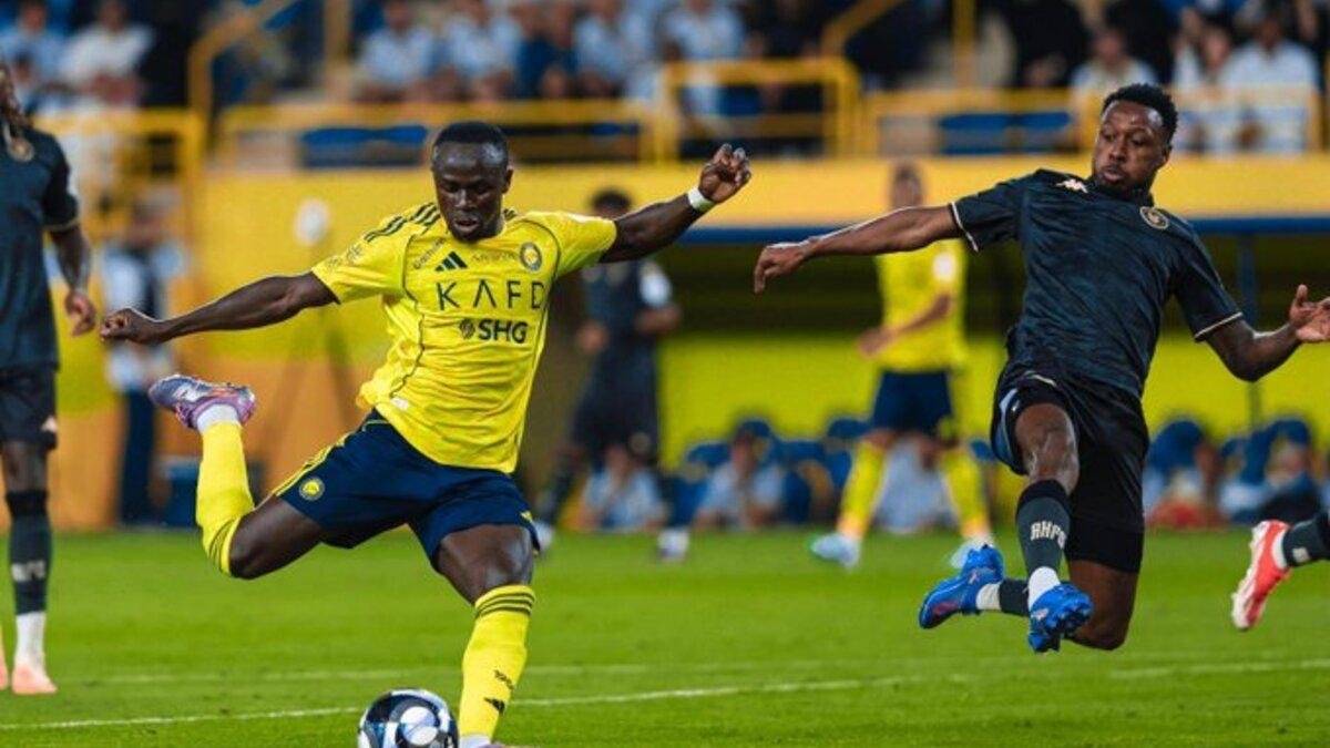 Two soccer players in a stadium duel for the ball; the yellow-kit player kicks while a dark-kit opponent closes in on the right. Two soccer players in a stadium duel for the ball; the yellow-kit player kicks while a dark-kit opponent closes in on the right.