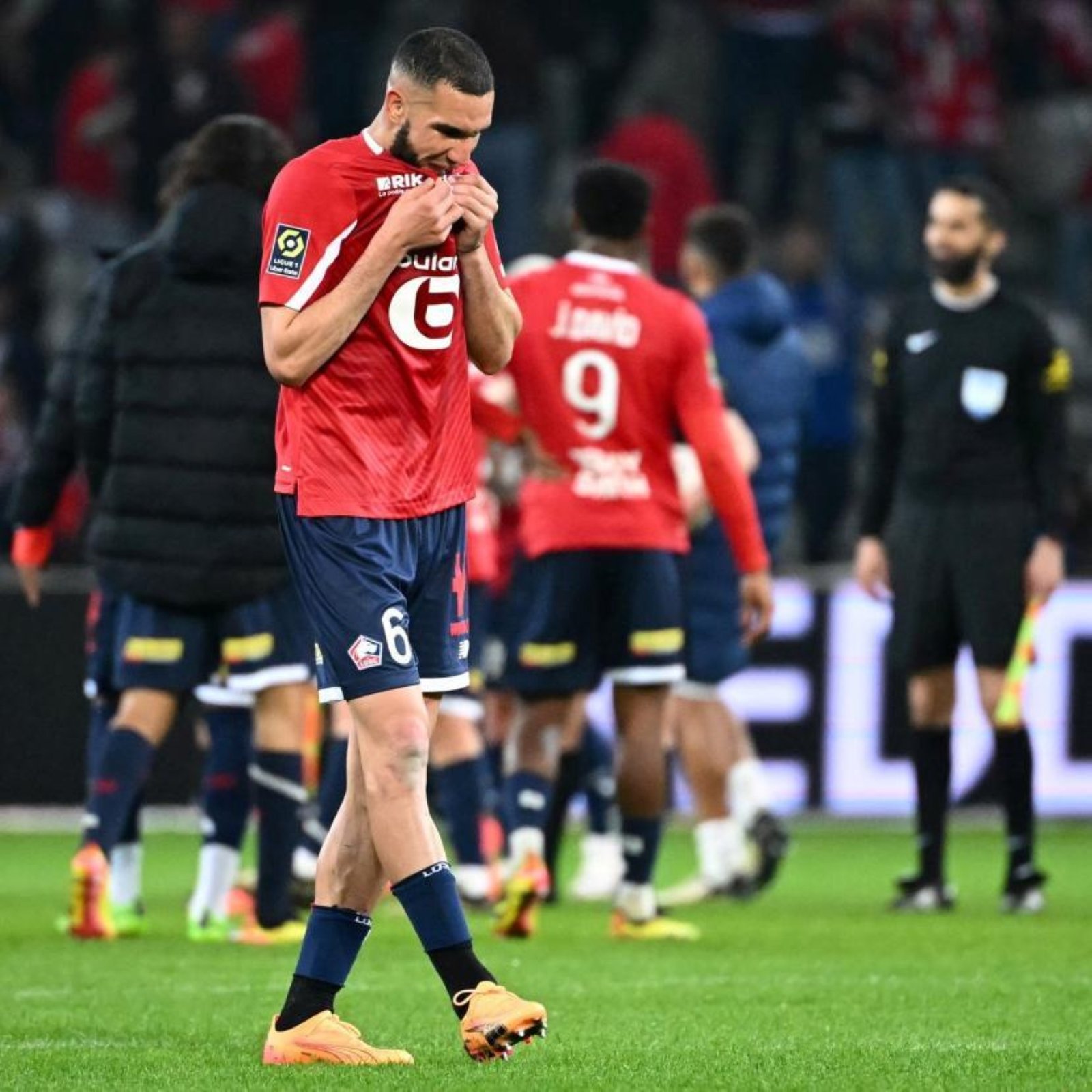 Soccer player in a red kit fixes his jersey on a green pitch, with teammates and spectators blurred in the background. Soccer player in a red kit fixes his jersey on a green pitch, with teammates and spectators blurred in the background.