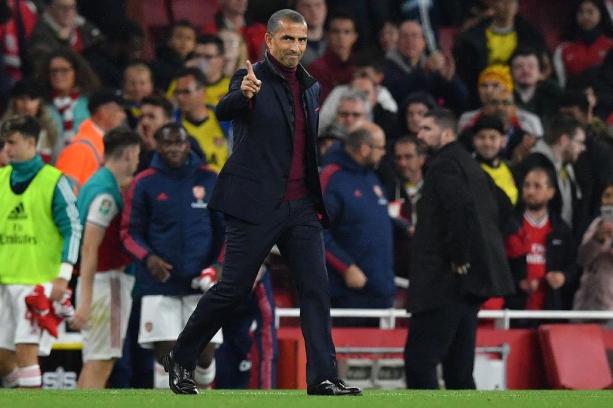 Football manager in a dark suit walks on the pitch, pointing with his right index finger as fans watch.