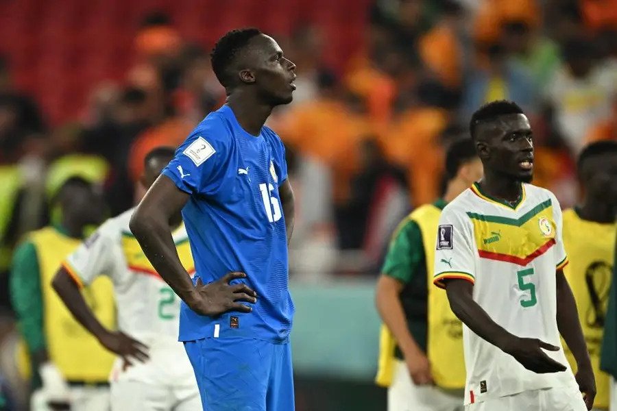 Soccer players on the field, blue jersey #16 stands with hands on hips while another player in white #5 looks on; blurred crowd in background.
