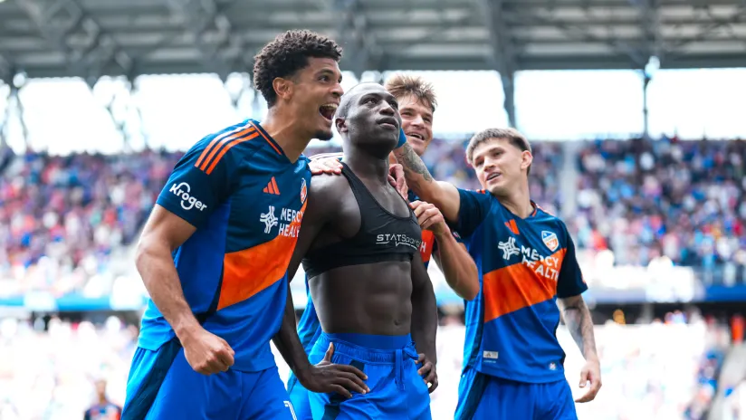 Soccer players celebrate a goal on the field with teammates surrounding a central player as the crowd cheers in the stands at a stadium game.