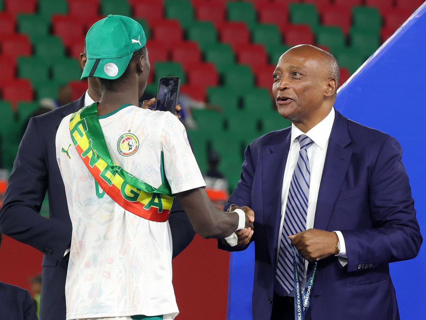 Soccer player wearing a white Senegal jersey with a green-red-yellow sash, green cap, shakes hands with a suited official wearing a blue suit in a stadium with red and green seats. Soccer player wearing a white Senegal jersey with a green-red-yellow sash, green cap, shakes hands with a suited official wearing a blue suit in a stadium with red and green seats. Soccer player wearing a white Senegal jersey with a green-red-yellow sash, green cap, shakes hands with a suited official wearing a blue suit in a stadium with red and green seats. Soccer player wearing a white Senegal jersey with a green-red-yellow sash, green cap, shakes hands with a suited official wearing a blue suit in a stadium with red and green seats.