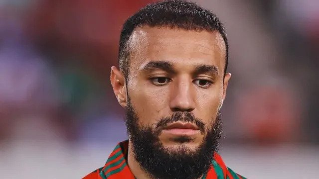 Close-up portrait of a man with a trimmed beard and short hair wearing a red-and-green sports jersey, looking downward with a pensive expression.