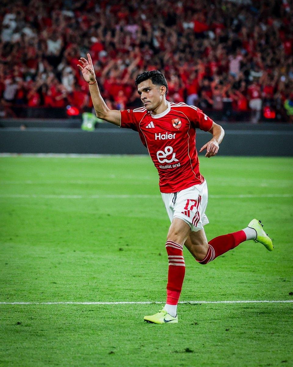 Soccer player in a red jersey celebrates a goal, arm raised, running on a green pitch with a cheering crowd behind.