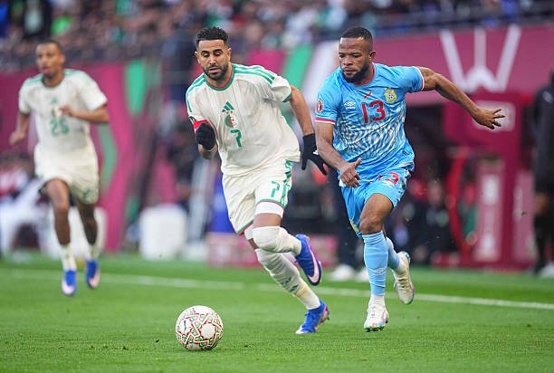 Two soccer players chase the ball on a green field during a match, one in white-and-green and the other in light blue, with a defender in white further back