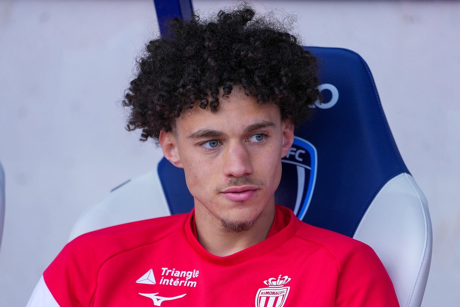 Young man with curly hair in a red jersey sits on a blue stadium seat, looking to the side with a calm expression. Young man with curly hair in a red jersey sits on a blue stadium seat, looking to the side with a calm expression.