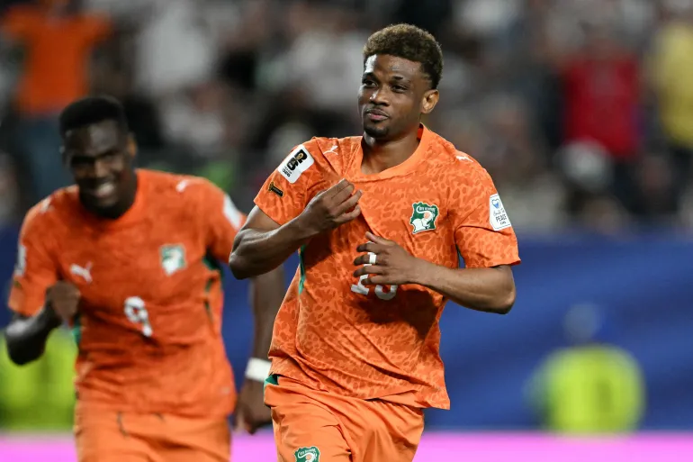 Two soccer players in orange uniforms run on the field during a match, with a blurred crowd behind them.