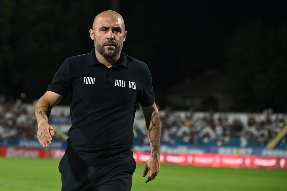 Bald man with a beard in a black polo shirt labeled 'TONY POLI IASI' walking on a soccer field at night with a blurred crowd behind him.