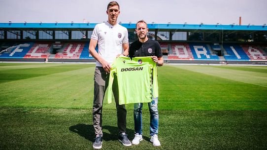 Two men on a soccer pitch hold up a bright neon green DOOSAN jersey for a jersey presentation on the field, stadium seating in the background.