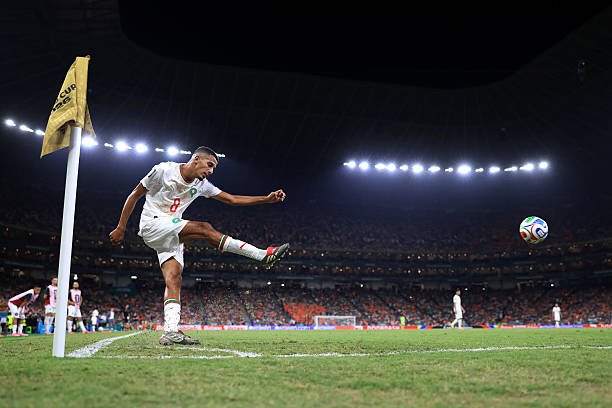 A footballer in a white kit kicks the ball from the corner flag as a large crowd watches a nighttime match behind him. A footballer in a white kit kicks the ball from the corner flag as a large crowd watches a nighttime match behind him.