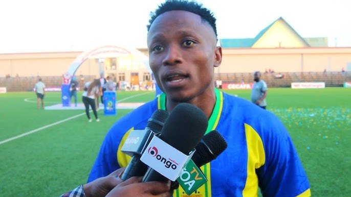 Male soccer player in a blue and yellow jersey being interviewed on a grassy football field, microphones held up to him by reporters.