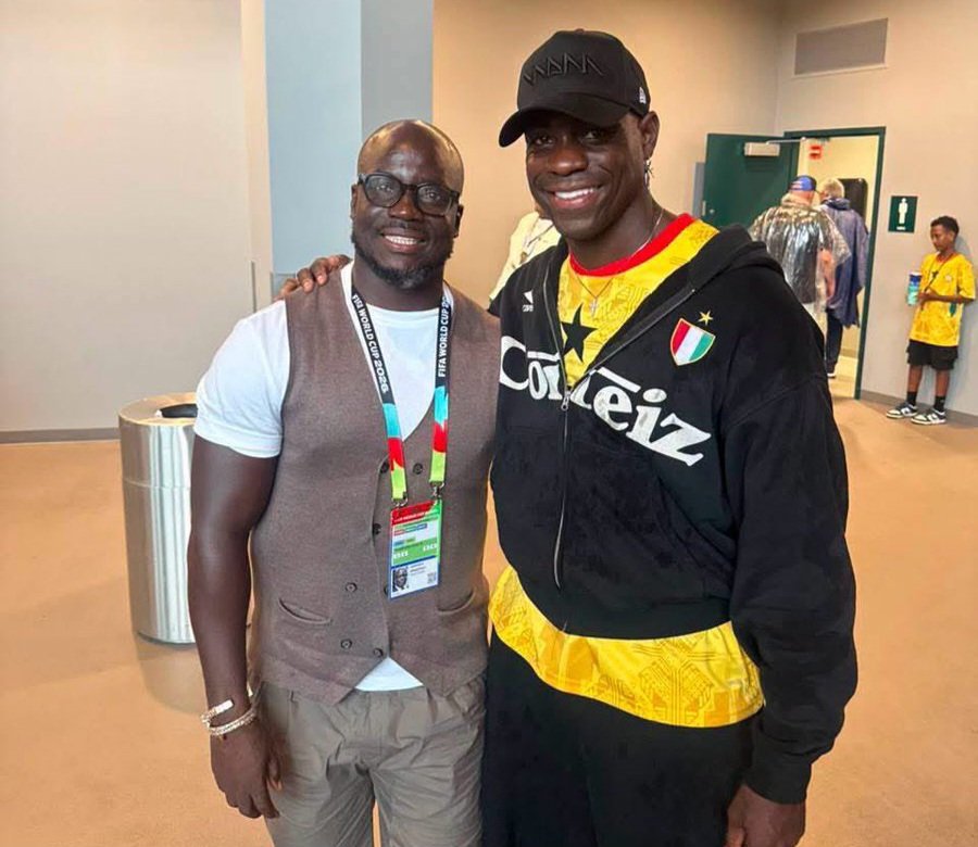 Two men pose together indoors, smiling for a photo at an event; one wears a beige vest, white shirt, and glasses, the other a black jacket and yellow shirt with a cap. Two men pose together indoors, smiling for a photo at an event; one wears a beige vest, white shirt, and glasses, the other a black jacket and yellow shirt with a cap.