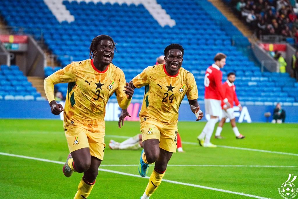 Two Ghanaian footballers in yellow kits celebrate on a green pitch, blue stadium seats behind them. Two Ghanaian footballers in yellow kits celebrate on a green pitch, blue stadium seats behind them.