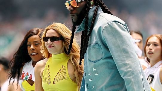 Group of celebrities walking at an outdoor event; a woman in neon yellow top and sunglasses beside a man with long braids and a light denim jacket.