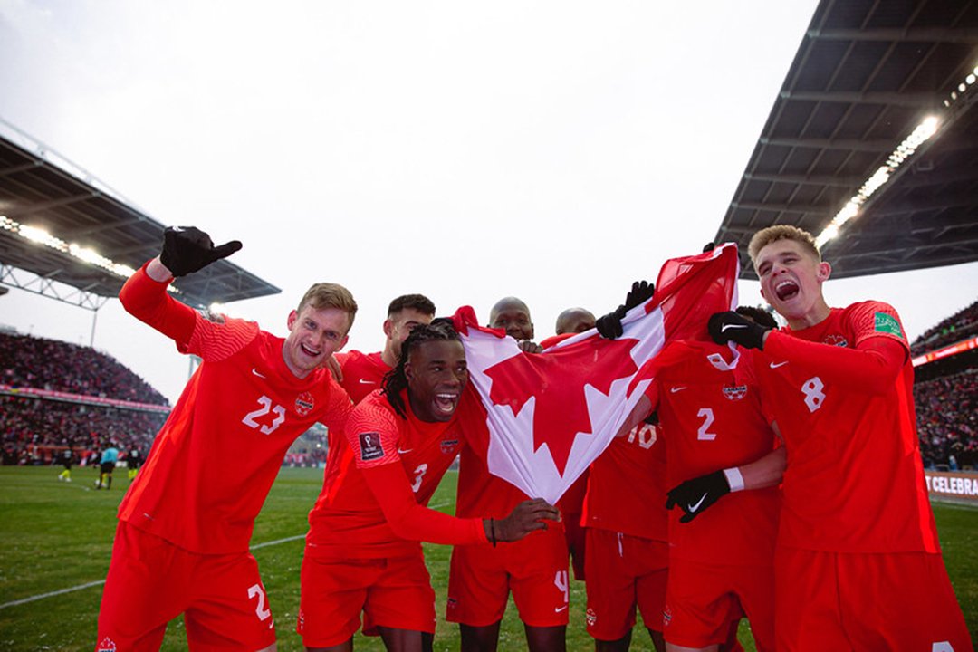 Canadian men’s soccer team celebrates a victory, holding a large Canadian flag on the field.