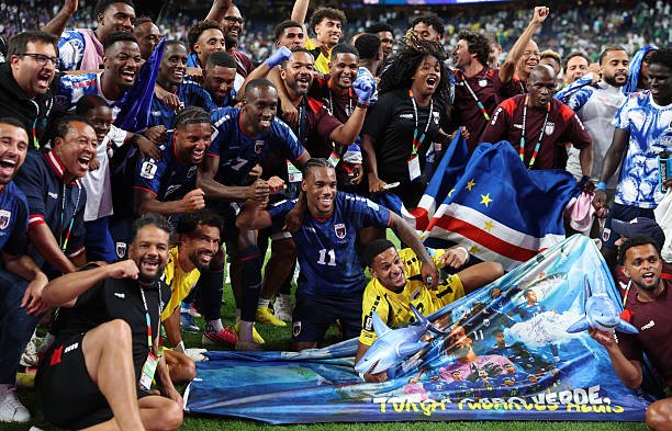 Diverse soccer players celebrate on the field, posing around a large blue banner on the ground as fans cheer in the stands. Diverse soccer players celebrate on the field, posing around a large blue banner on the ground as fans cheer in the stands.