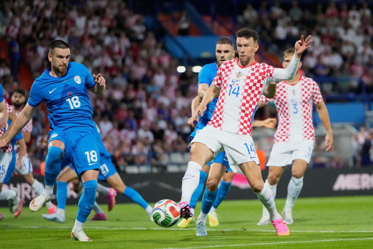 Soccer scene: Croatia player in white-and-red checkered kit swings a leg toward the ball as blue-clad defenders approach.