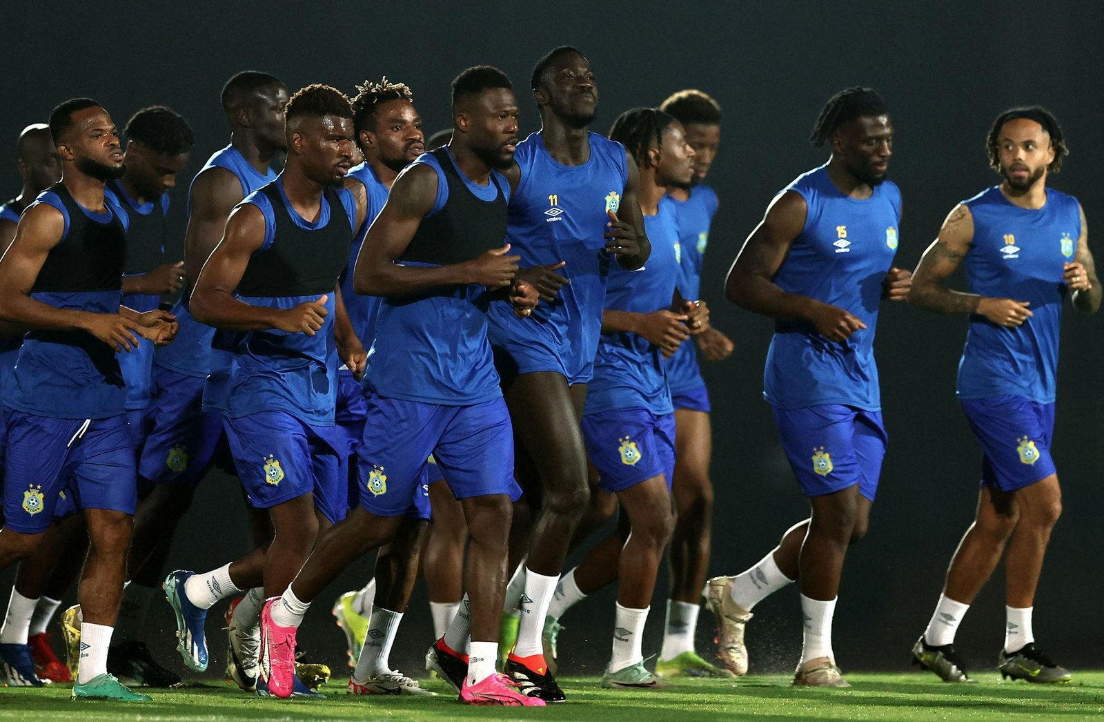 Group of male soccer players in blue training kits running together on a field, silhouettes against a dark background.