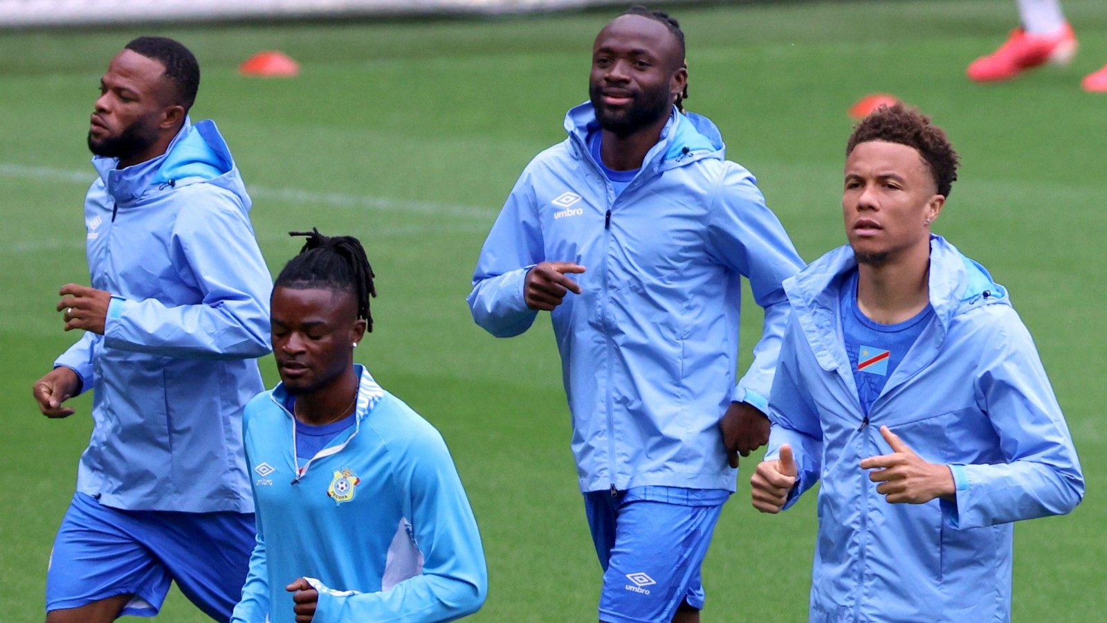 Soccer players in light-blue jackets jogging on a grassy training field.