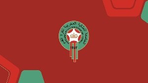 Circular green logo with Arabic inscription around the border and a central torch motif, set on a red background with geometric shapes. Circular green logo with Arabic inscription around the border and a central torch motif, set on a red background with geometric shapes.